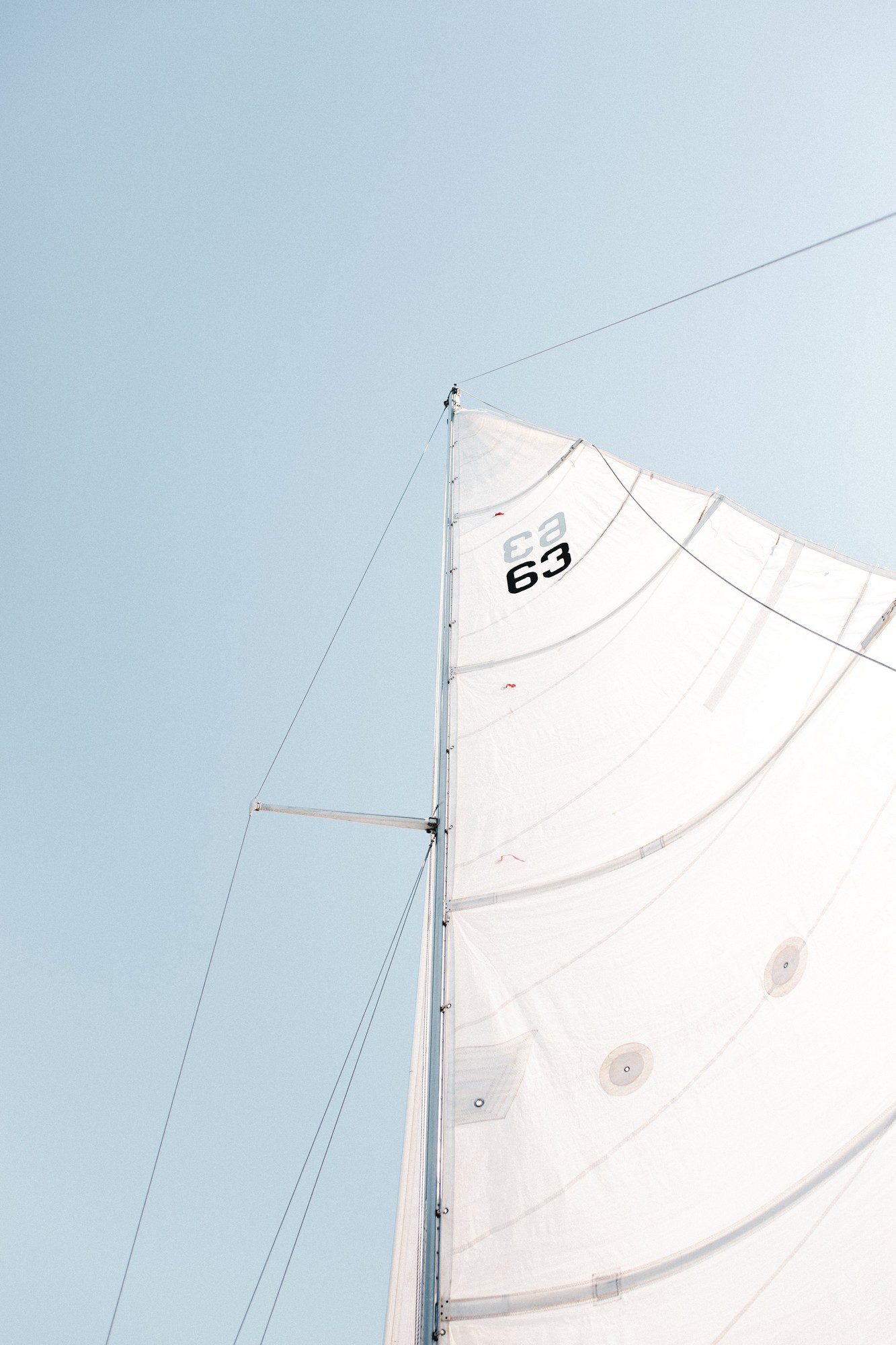 The image displays a close-up of a white sail on a sailing boat, with a clear blue sky in the background. The sail has the number "63" printed on it and is attached to a mast on the left side. Visible are the ropes and other hardware associated with adjusting and securing the sail.