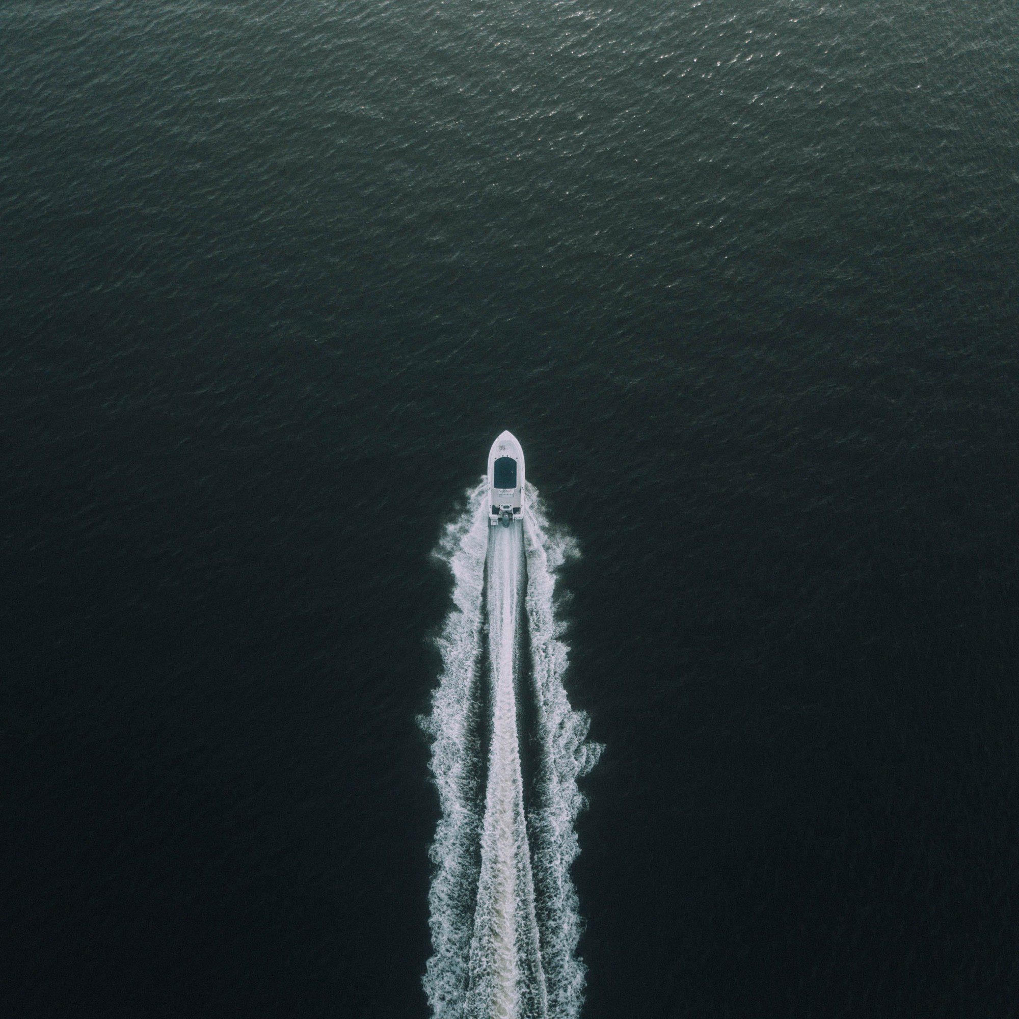 This image shows an aerial view of a boat moving through water. The boat is creating a distinct wake behind it, characterized by white, frothy waves on either side of its path. The water appears dark, which contrasts with the white wake. This perspective gives a sense of the boat's speed and direction as it travels across the water's surface.