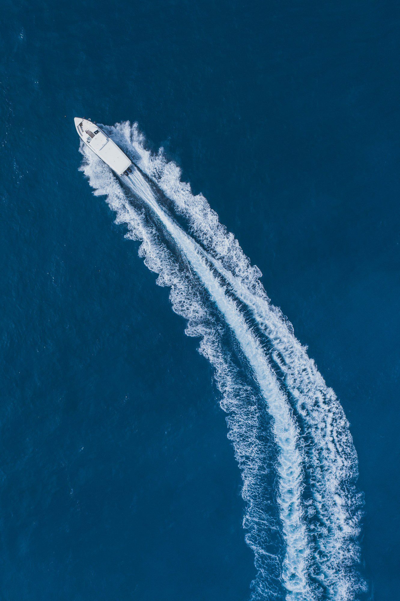 This is an aerial image of a speedboat moving through a body of water, most likely the ocean, given the intense blue coloration. The boat is generating a wake pattern behind it, with white frothy water fanning out on either side due to its high speed. The overhead perspective creates a pleasing visual contrast between the deep blue of the water and the white of the wake.