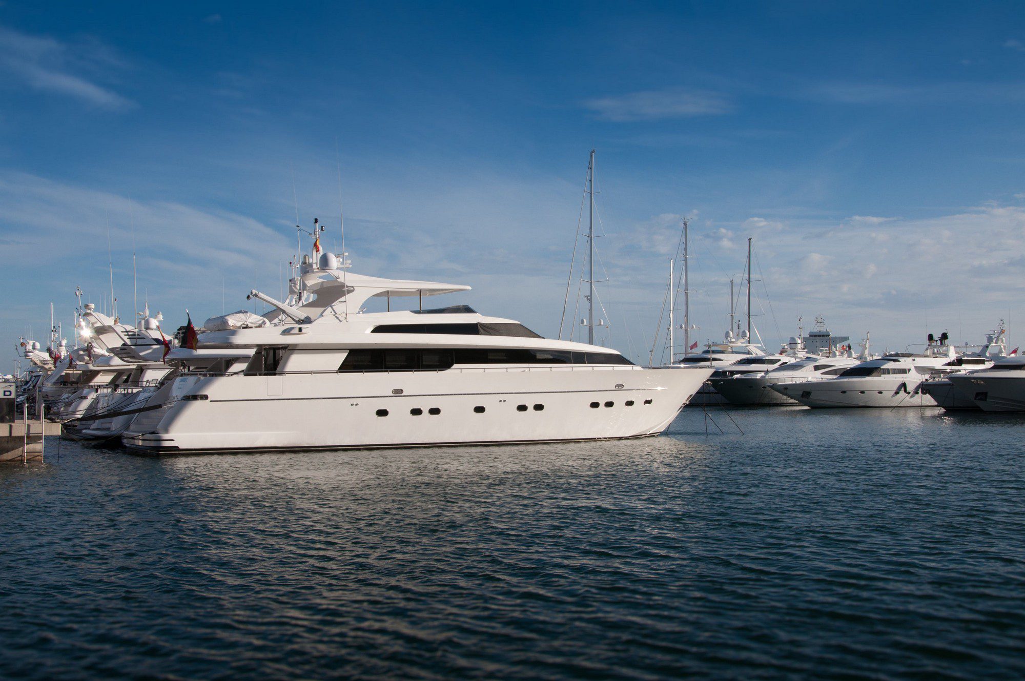 The image shows a marina with several luxury yachts moored in calm water. The yachts vary in size and design but are predominantly white. The sky is clear with a few clouds, indicating fair weather conditions. No people are distinctly visible, maintaining privacy and focusing the viewer's attention on the luxury vessels and tranquil marine setting.