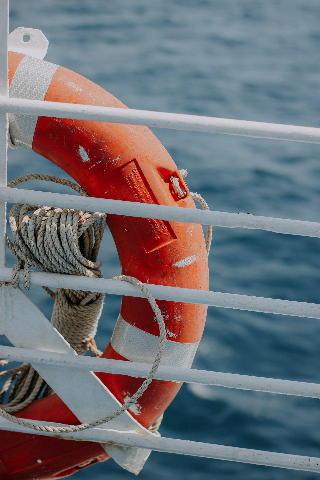 The image shows a lifebuoy attached to the railings of what appears to be a boat or ship. The lifebuoy is orange with reflective material and is tied with a rope, suggesting that it is ready for use in case of an emergency. In the background, there is a body of water which could be the sea or an ocean, indicating that the vessel is on the water. The focus is mainly on the lifebuoy, and it illustrates standard safety equipment found on maritime vessels.