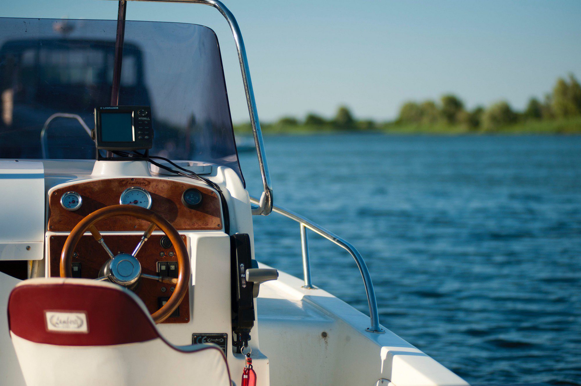 This image shows the helm of a boat with a clear view of the steering wheel and the control panel. The steering wheel is made of wood, and behind it, there's what appears to be a dashboard with various gauges and switches, featuring a wood panel finish as well. Mounted on top of the dashboard is a small electronic device, which could be a fish finder or a GPS navigation unit, labeled "LOWRANCE." In front of the wheel, there's a windscreen to protect the driver from the elements, and part of the boat's seating can be seen with the brand name "Cobalt" stitched on the backrest. The backdrop of the photo is a body of water, suggesting that the boat is on a river or lake, and there's foliage visible on the distant shoreline under a clear blue sky.
