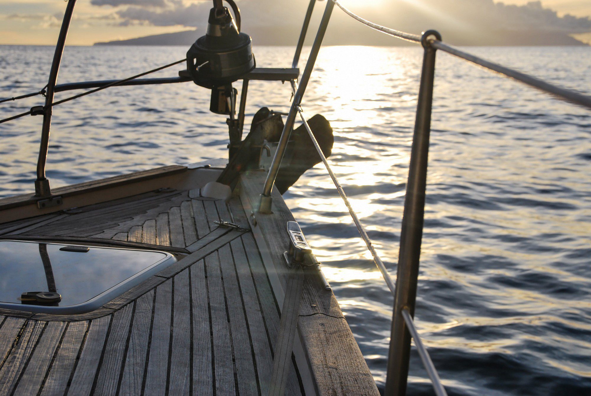 The image shows a view from the deck of a sailing boat during what appears to be either sunrise or sunset, judging by the warm light and low angle of the sun reflected on the water. The focus is on the wooden deck, which features various nautical elements like cleats, a winch, a bow pulpit, stainless steel railings, and rigging lines. The calm sea and the soft glow of the sun create a tranquil and inviting maritime scene. There is land visible on the horizon, suggesting the boat isn't far from shore.