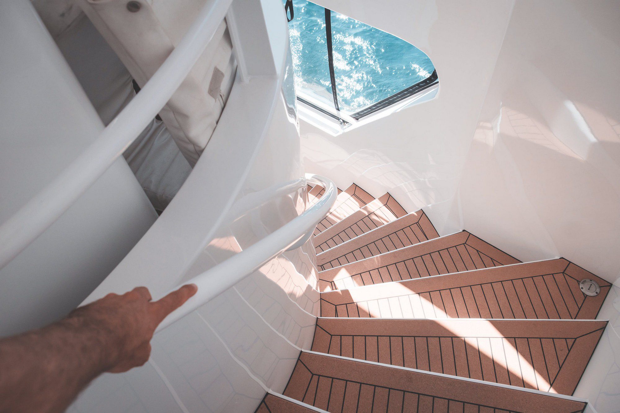 The image shows a staircase leading down inside a modern-looking structure, likely a boat or yacht, as suggested by the window offering a view of the water and the boat's structure. The stairs have brown non-slip treads and white walls, with a handrail on one side for safety. The interior is well-lit, possibly by natural light coming from above. There's a person's arm visible, pointing at something outside the window, likely indicating something of interest in the water or in the distance. The setting gives the impression of a luxury or recreational vessel.